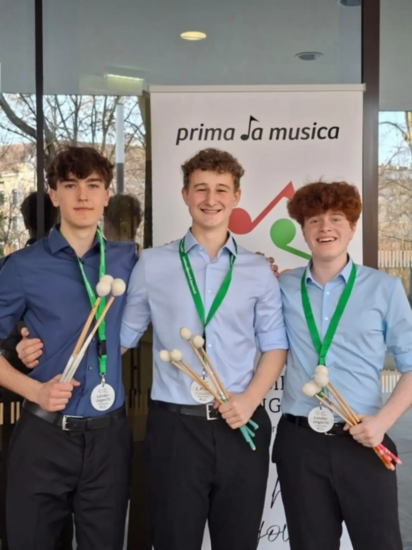 Three young men in blue shirts and black pants stand next to each other, each holding a set of drumsticks. They are standing in front of a white banner with the text 'prima Ja musica'.