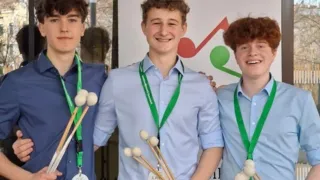 Three young men in blue shirts and black pants stand next to each other, each holding a set of drumsticks. They are standing in front of a white banner with the text 'prima Ja musica'.