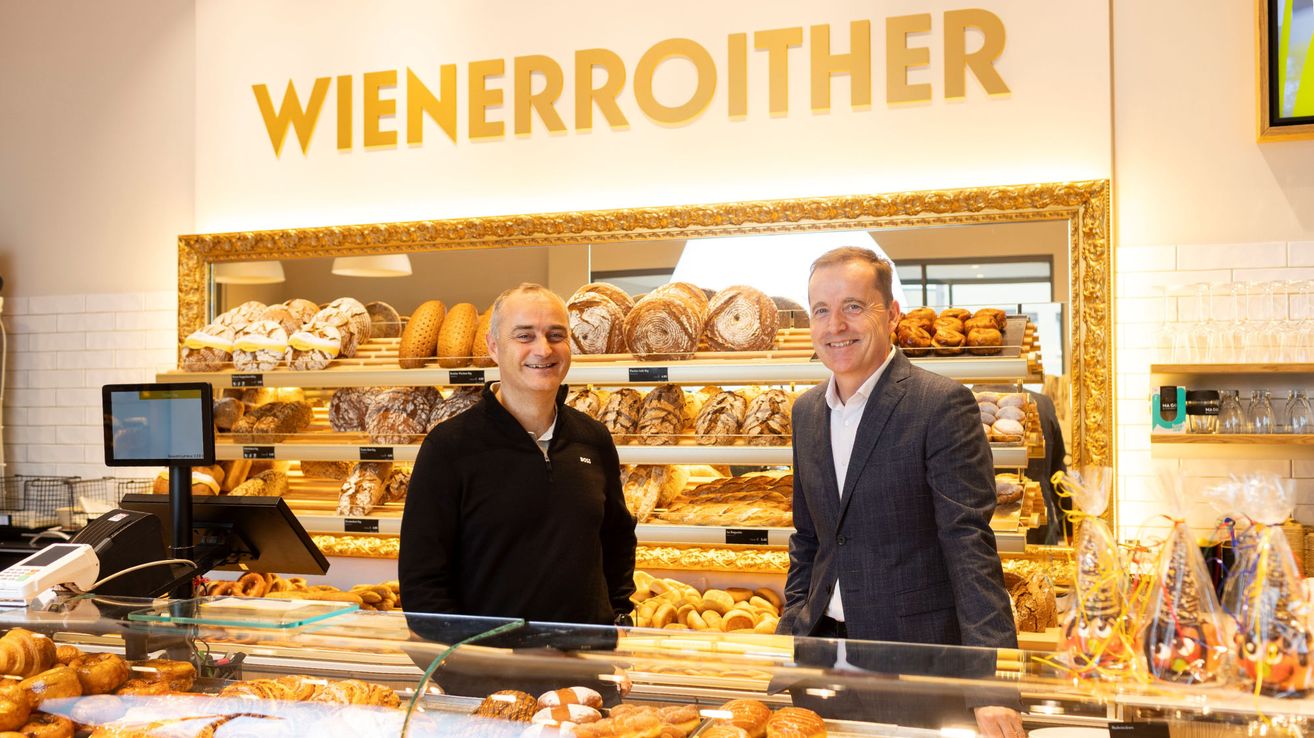 Zwei Männer stehen lächelnd hinter einer Theke in einer Bäckerei und posieren für ein Foto. Das Schild darüber lautet 'Wienerroither'. Dahinter sind Regale mit verschiedenen Brotsorten und Gebäck gefüllt.