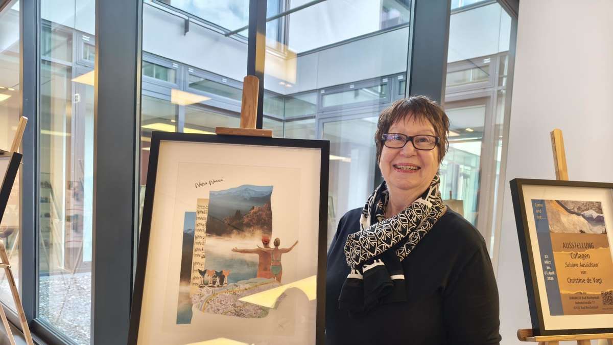 A woman stands next to a framed art piece with a scenic collage, smiling and wearing glasses and a scarf. Behind her is a glass wall reflecting a modern building.