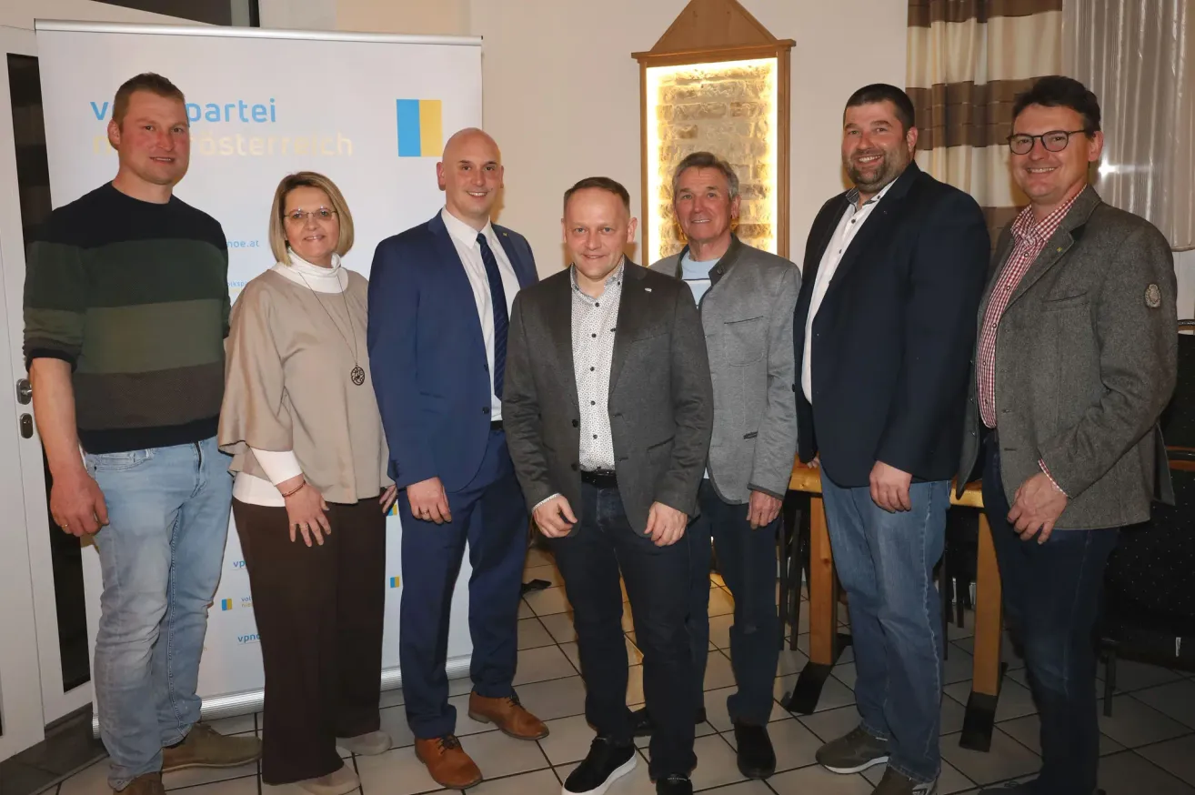 Six people stand in a room. Two women and four men. The woman wears glasses, a necklace, and a bracelet. The men wear suits, ties, and leather shoes. A wooden desk is behind them.
