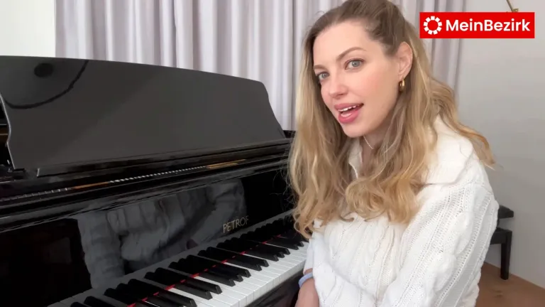 Eine Frau mit blonden Haaren sitzt vor einem schwarzen Petrof-Klavier, lächelt und schaut direkt in die Kamera.