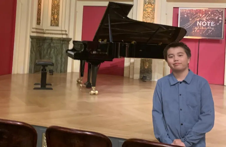 A young boy stands next to a grand piano on a stage, wearing a blue shirt and posing for a photo. The piano is a Steinway model, and there are two chairs in front of him. The stage is wooden with a red backdrop.