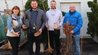 Four individuals stand in front of a white building, each holding a plant. The man on the right wears a blue jacket, while the man next to him wears a white hoodie. The two women on the left wear denim jackets.