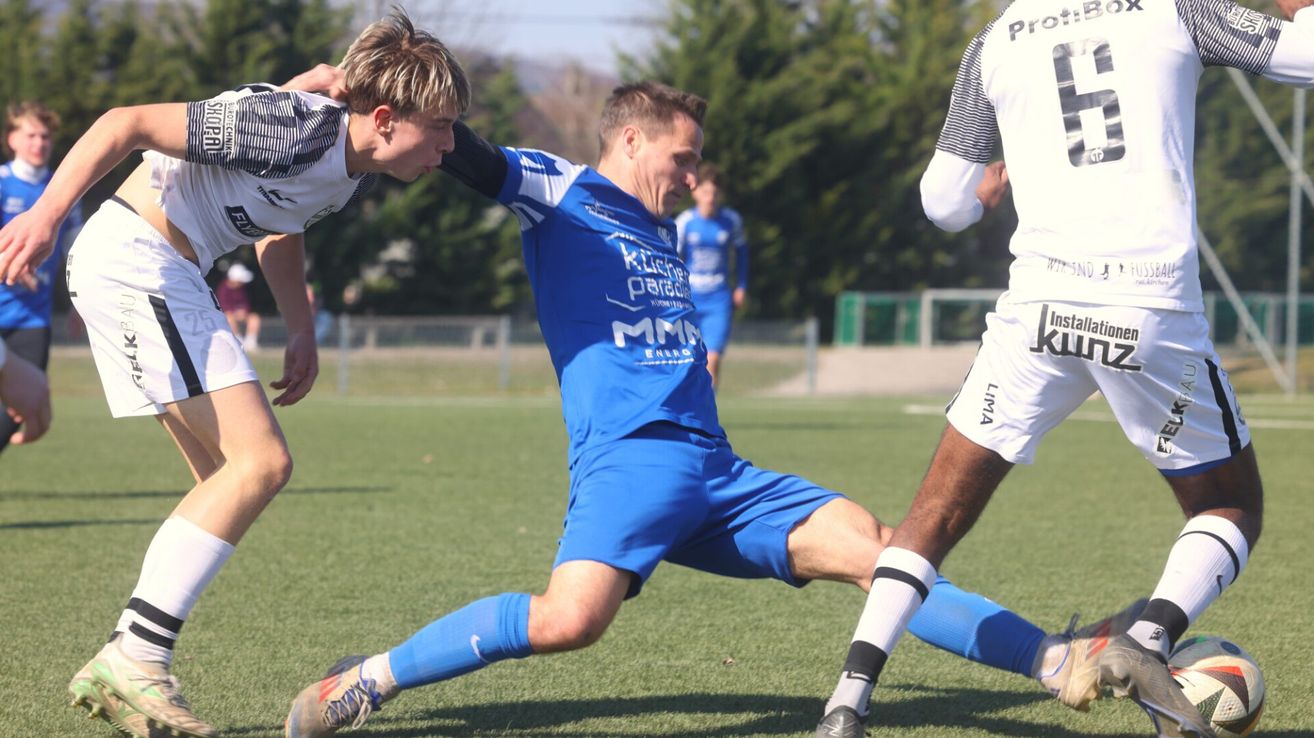 Ein Fußballspieler in Blau verteidigt gegen einen Gegner in Weiß. Beide sind auf einem Feld mit Bäumen und einem Zaun im Hintergrund.