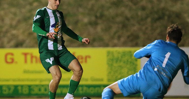 Ein Fußballspieler in einem grünen Trikot ist in Bewegung, möglicherweise beim Balltreten, während ein anderer Spieler in Blau in der Nähe ist, beide auf einem Feld mit einem gelben Banner im Hintergrund.