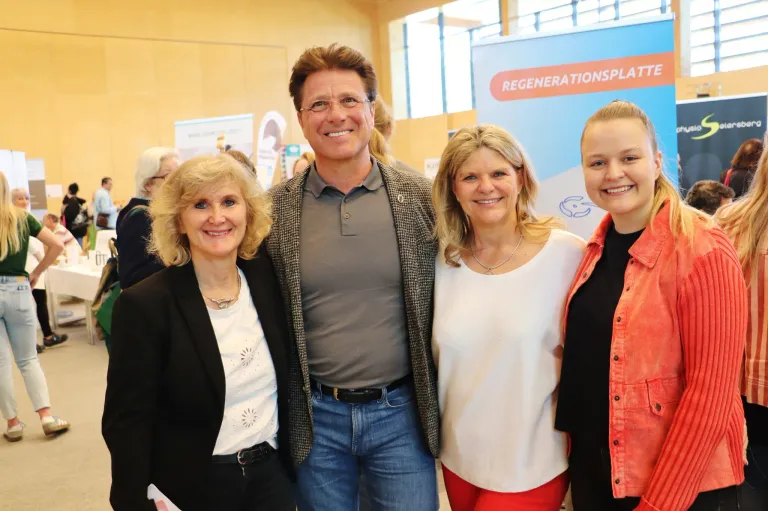 Eine Gruppe von vier Personen steht zusammen und lächelt für ein Foto vor einem Schild mit dem Wort 'Regenerationsplatz.' Der Mann in der Mitte trägt eine Brille und ein graues Hemd, während die anderen Jacken tragen.