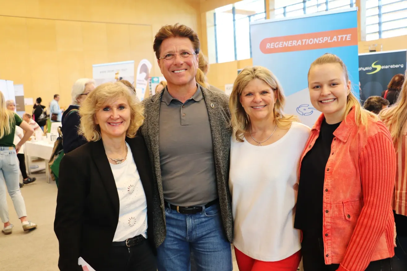 Eine Gruppe von vier Personen steht zusammen und lächelt für ein Foto vor einem Schild mit dem Wort 'Regenerationsplatz.' Der Mann in der Mitte trägt eine Brille und ein graues Hemd, während die anderen Jacken tragen.