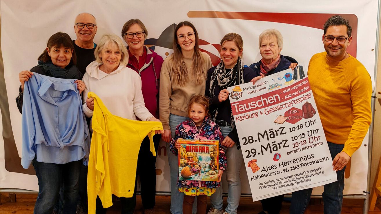 Eine Gruppe von Erwachsenen und Kindern posiert zusammen mit einem Banner auf Deutsch. Ein junges Mädchen hält eine Box und eine Frau hält ein Banner. Das Banner enthält Details zu einer Kleidungstausch-Veranstaltung.