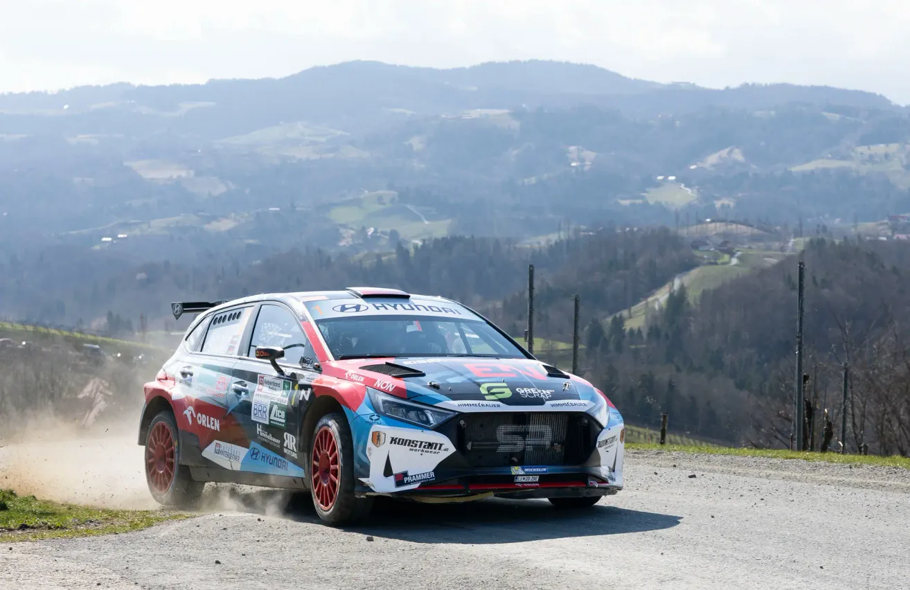 Ein Rallyeauto mit Hyundai-Marke fährt auf einer kurvenreichen Bergstraße, umgeben von üppigen grünen Bäumen und Bergen.