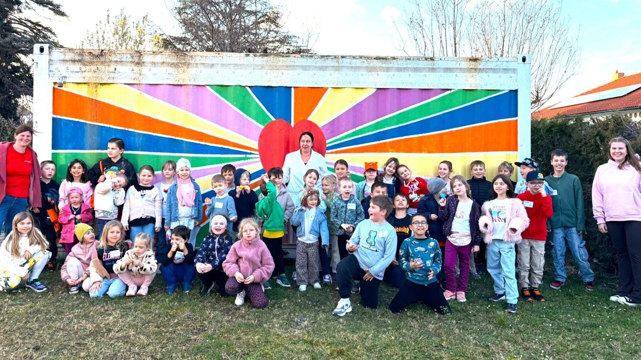 Eine Gruppe von Kindern in Winterkleidung posiert vor einer farbenfrohen Wandmalerei mit einem großen roten Herzen und einer Frau, die hinter ihnen auf einem Rasenfeld steht.