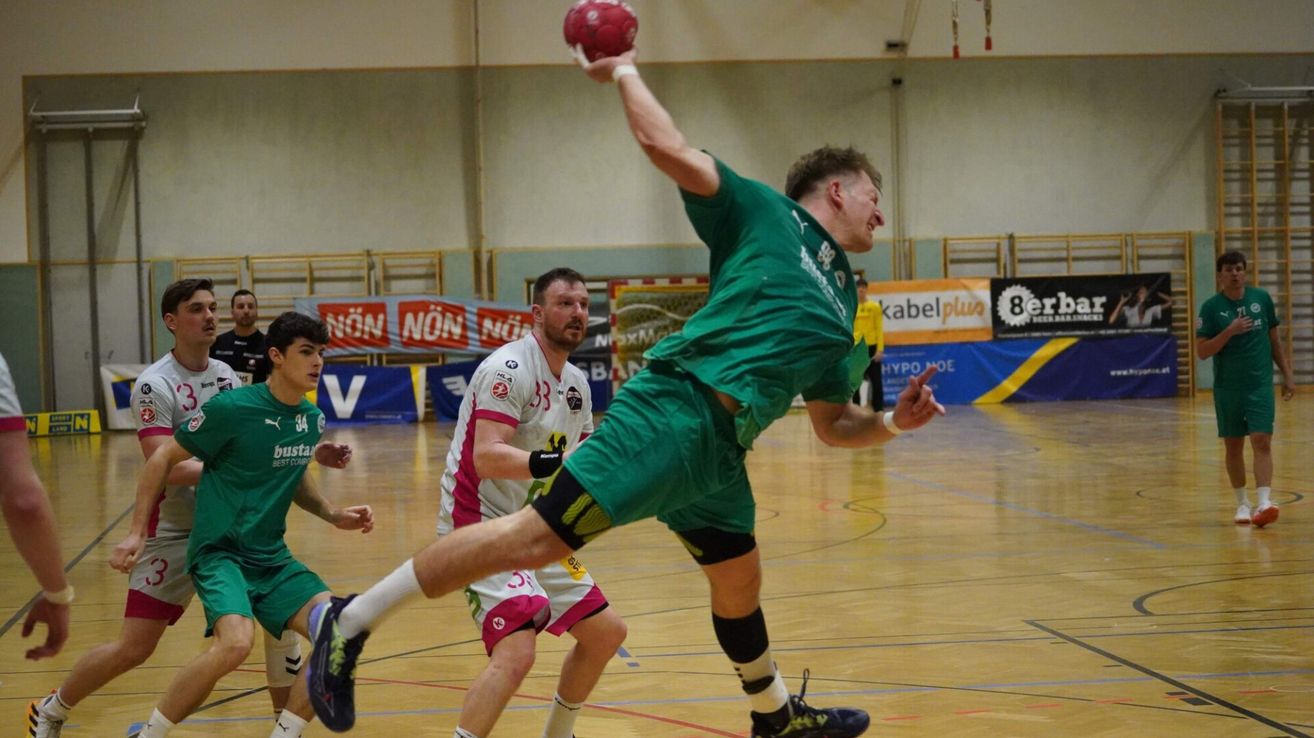 Ein Mann in einem grünen Trikot ist in der Luft, hält einen roten Ball und scheint ihn zu werfen. Hinter ihm steht ein anderer Mann in einem weißen Trikot. Sie sind auf einem überdachten Basketballplatz.