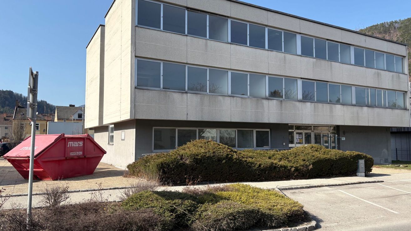 Ein großes, graues Gebäude mit mehreren Fenstern, möglicherweise ein Bürogebäude, hat einen roten Müllcontainer links. Vor dem Gebäude stehen Sträucher.