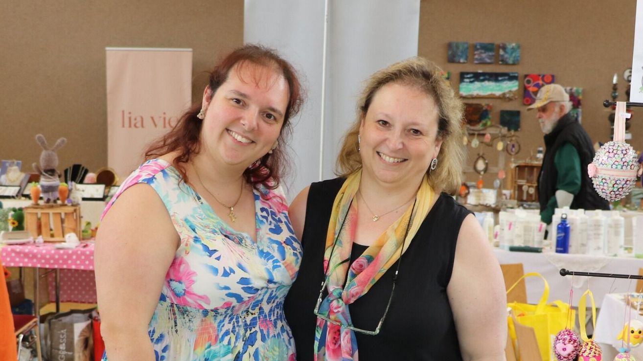 Zwei Frauen stehen nebeneinander, lächelnd, in einem Raum mit einer Wand voller Kunstwerke. Die Frau auf der linken Seite trägt ein Blumenkleid und eine Halskette. Die Frau auf der rechten Seite trägt ein schwarzes ärmelloses Oberteil mit einem Blumenschal.