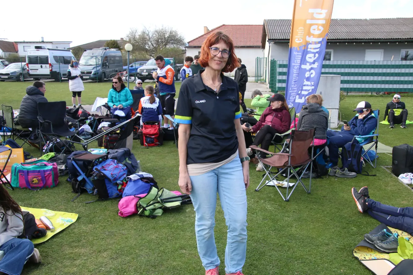 Eine Frau in einem schwarzen Shirt steht auf einem Rasenfeld. Mehrere Leute sitzen mit Taschen und Stühlen herum. Ein Banner ist im Hintergrund zu sehen.