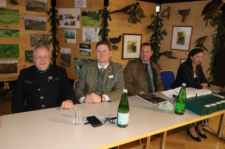 Drei Männer in formeller Kleidung sitzen an einem Tisch mit einem Buch und zwei Flaschen grüner Flüssigkeit. Die Wand hinter ihnen hat Bilder von Vögeln und Pflanzen.