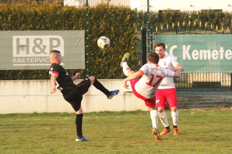 Drei Männer spielen Fußball auf einem Feld. Einer in Schwarz tritt den Ball, während zwei in Rot und Weiß versuchen, ihn zu blockieren. Dahinter steht eine Mauer mit einem Banner und dahinter ein Tor mit weiteren Bannern.