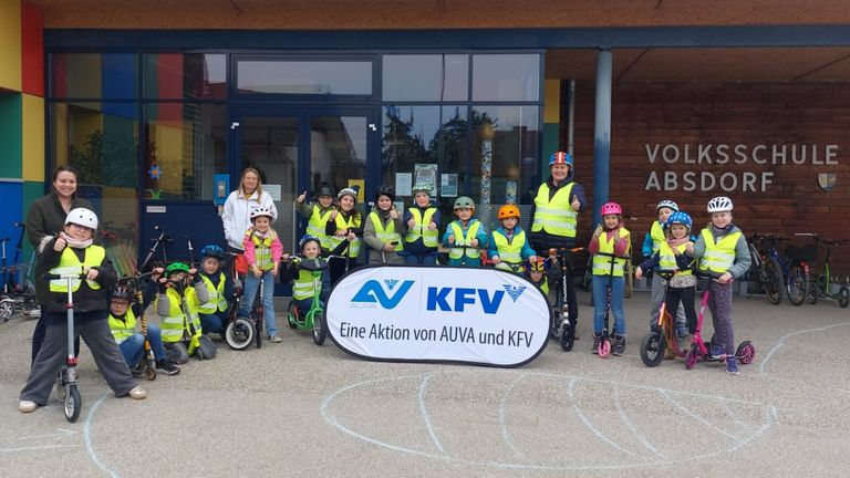 Eine Gruppe von Kindern und Erwachsenen in Sicherheitswesten posiert mit ihren Rollern vor einem Gebäude. Ein Banner mit dem Text 'AUVA KFV' steht in der Mitte.