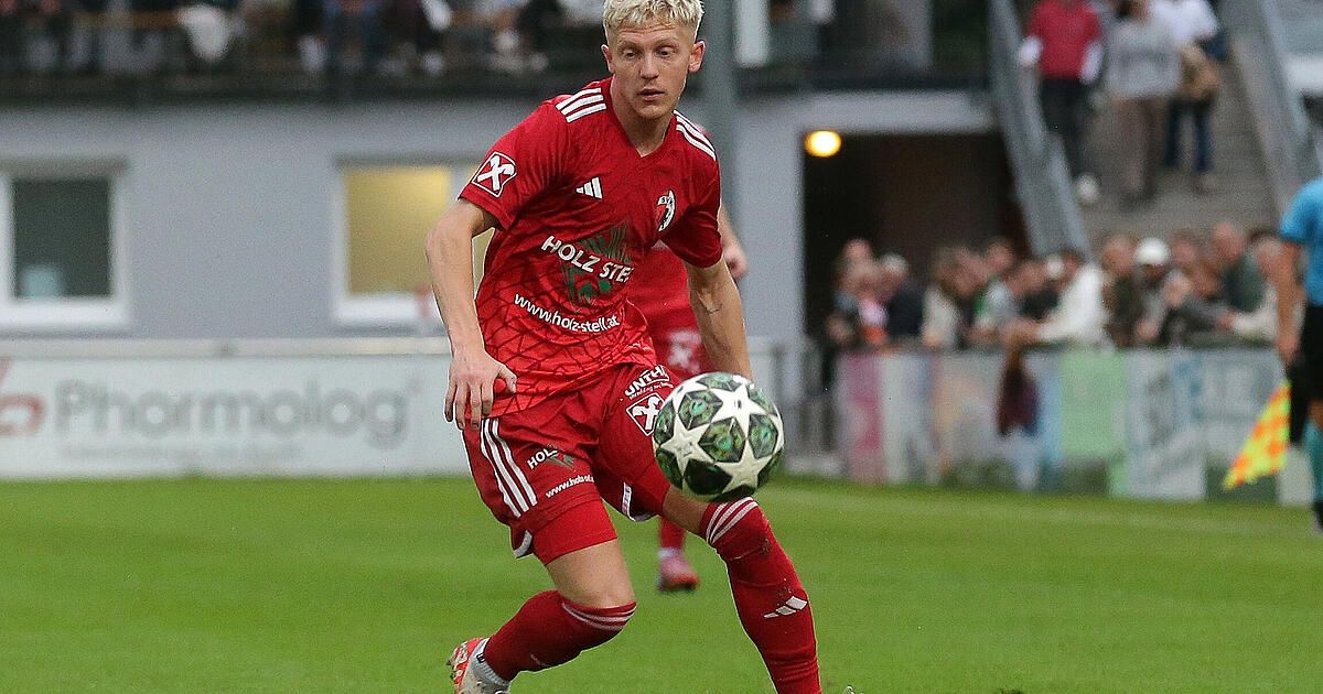 Ein Fußballspieler in einem roten Trikot tritt einen Ball auf dem Feld. Zuschauer sehen von den Tribünen zu.