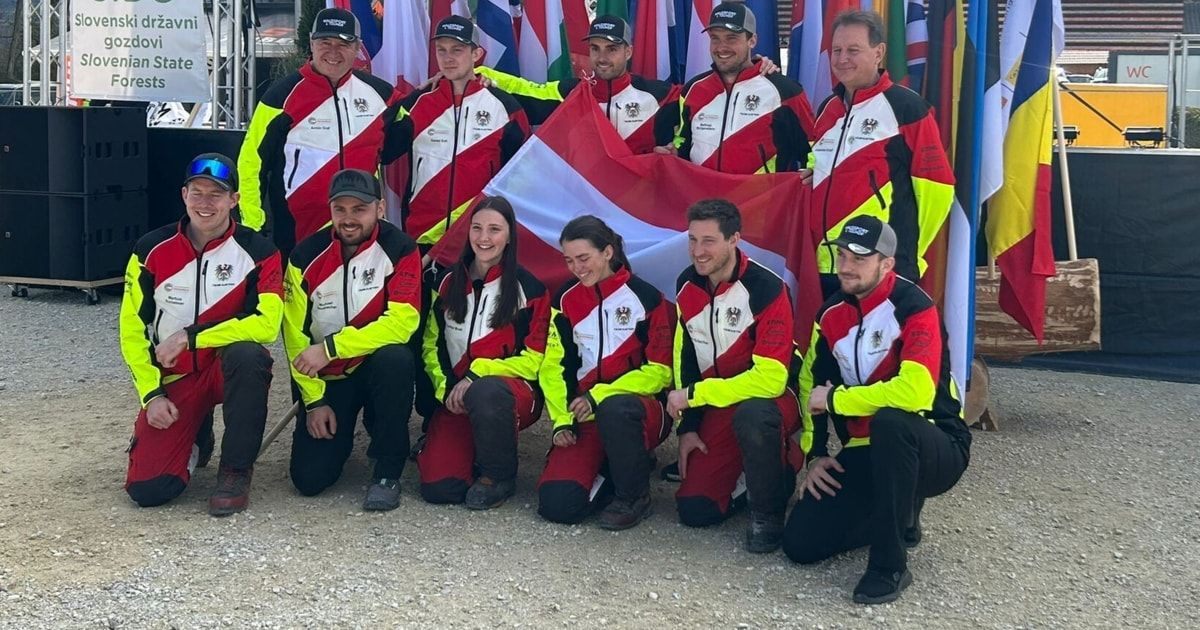 Ein Team von Rennfahrern in roten, weißen und schwarzen Uniformen, die eine Flagge halten, posiert für ein Foto. Dahinter sind mehrere Fahnen an Stangen befestigt.