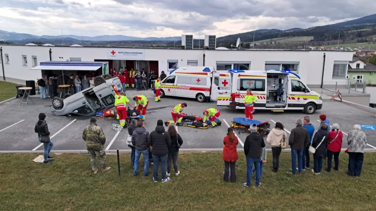 Rettungskräfte in gelben Uniformen versorgen Patienten vor einem Krankenwagen. In der Nähe liegt ein beschädigtes Fahrzeug auf der Seite. Zuschauer beobachten vom Gras aus.