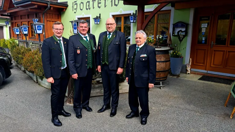 Vier Männer in Anzügen stehen vor einem Gebäude mit dem Namen 'Gasthaus'. Sie tragen grüne Westen und haben Medaillen an ihren Jacken. Hinter ihnen steht ein Fass, Pflanzen und ein Fenster mit Blick auf ein Auto.