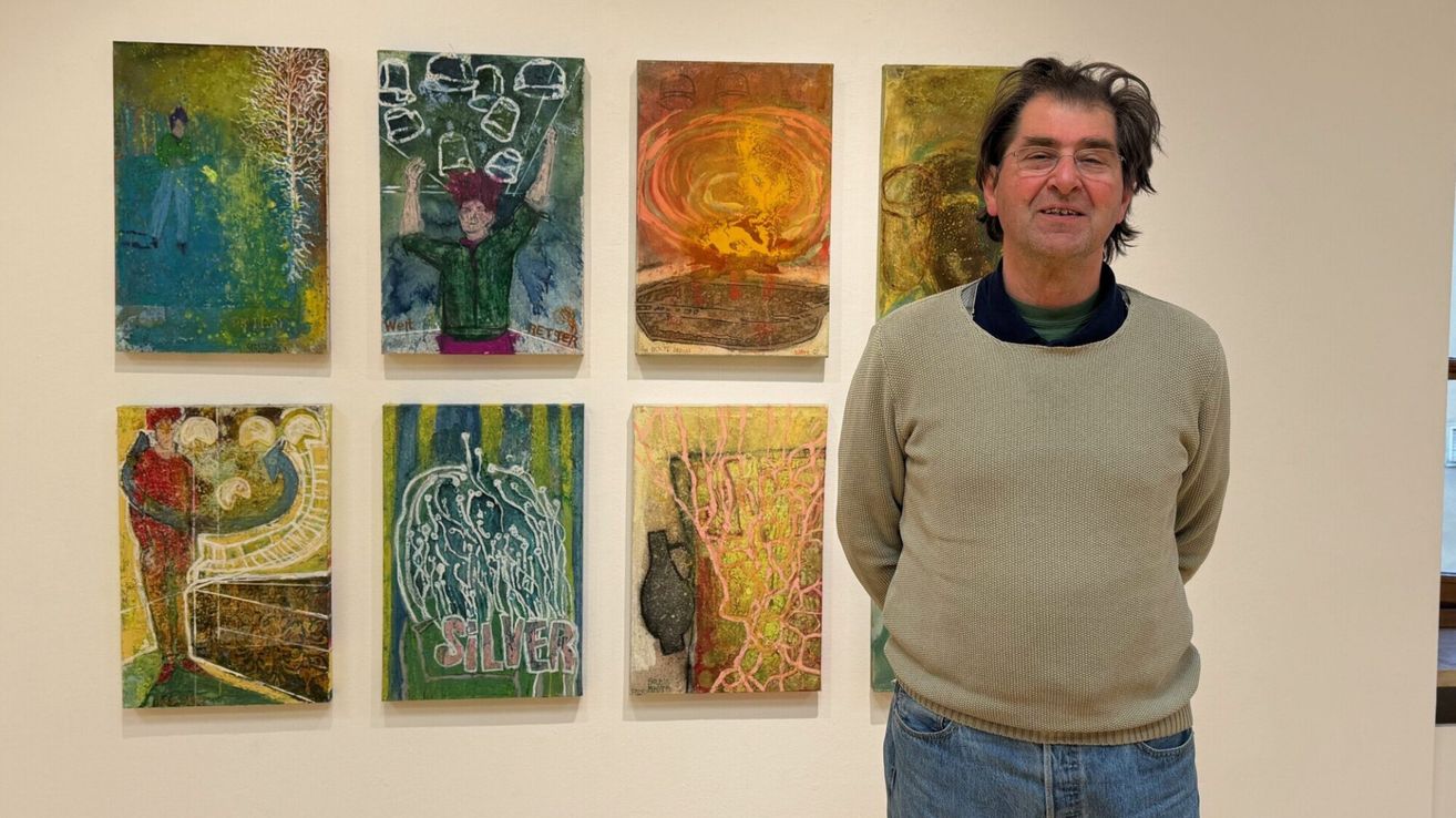 Ein Mann in Pullover und Jeans steht vor einer Wand mit mehreren Gemälden und lächelt für die Kamera.