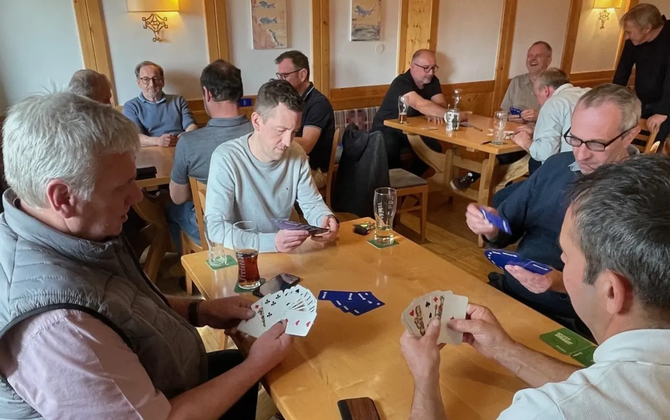 Mehrere Männer sitzen an einem Tisch in einer Kneipe. Sie halten Spielkarten und Gläser Bier in den Händen. Ein Mann schaut auf seine Karten. Der Tisch hat eine Bierdeckel und Untersetzer.