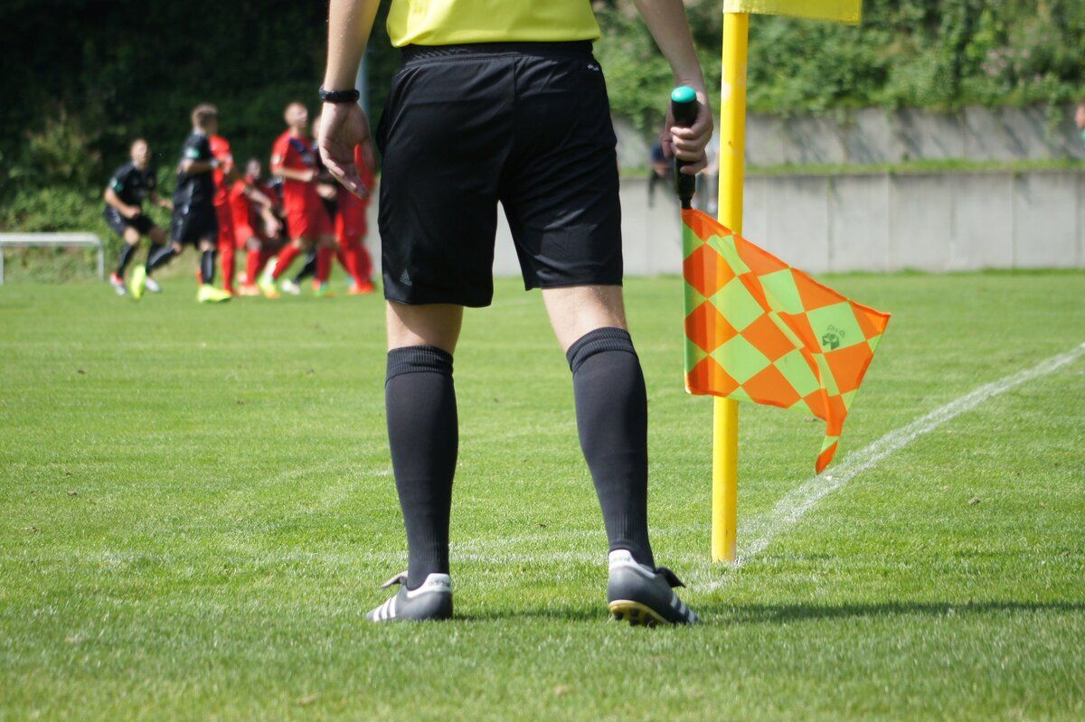 Ein Fußballschiedsrichter steht auf einem Feld mit Pfeife und Flagge, Spieler in roten Trikots im Hintergrund.