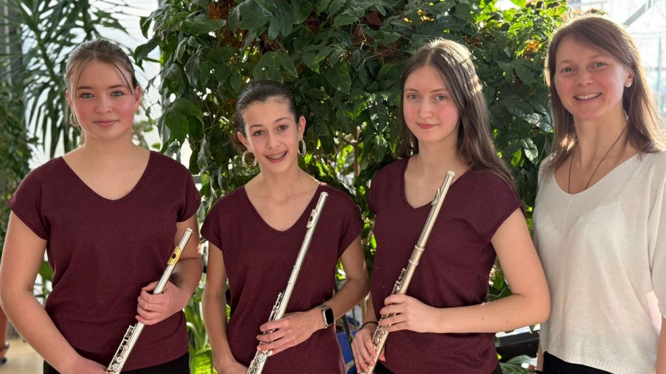 Drei junge Frauen, die rote T-Shirts tragen, lächeln und posieren für ein Foto, während sie Flöten halten. Ein grüner Busch ist hinter ihnen.