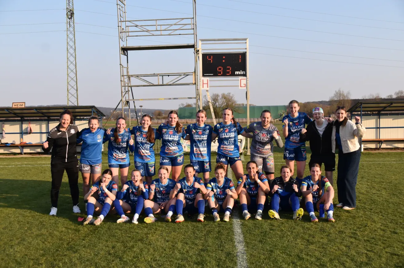 Eine Frauenfußballmannschaft posiert für ein Foto auf einem Spielfeld, wobei die Anzeigetafel 4:1 und 33 verbleibende Minuten im Spiel anzeigt.
