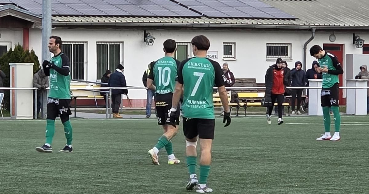 Zwei Fußballspieler laufen auf dem Feld, einer trägt das Trikot Nummer 10 und der andere 7. Dahinter steht ein Gebäude mit Solarpanelen und mehrere Personen.