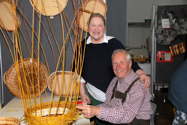 Ein älterer Mann in einem karierten Hemd und suspenders sitzt neben einer Frau in einem schwarzen Pullover. Sie lächeln beide und posieren für ein Foto vor einer gewebten Korbstruktur.