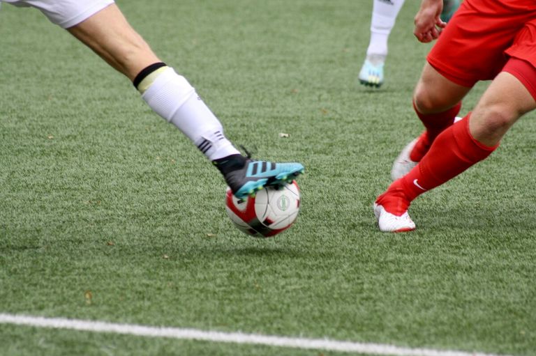 Ein Fußballspieler in weißen Uniformen tritt mit einem blauen Schuh einen Fußball auf einem grünen Feld. Ein anderer Spieler in roten Uniformen ist in der Nähe.