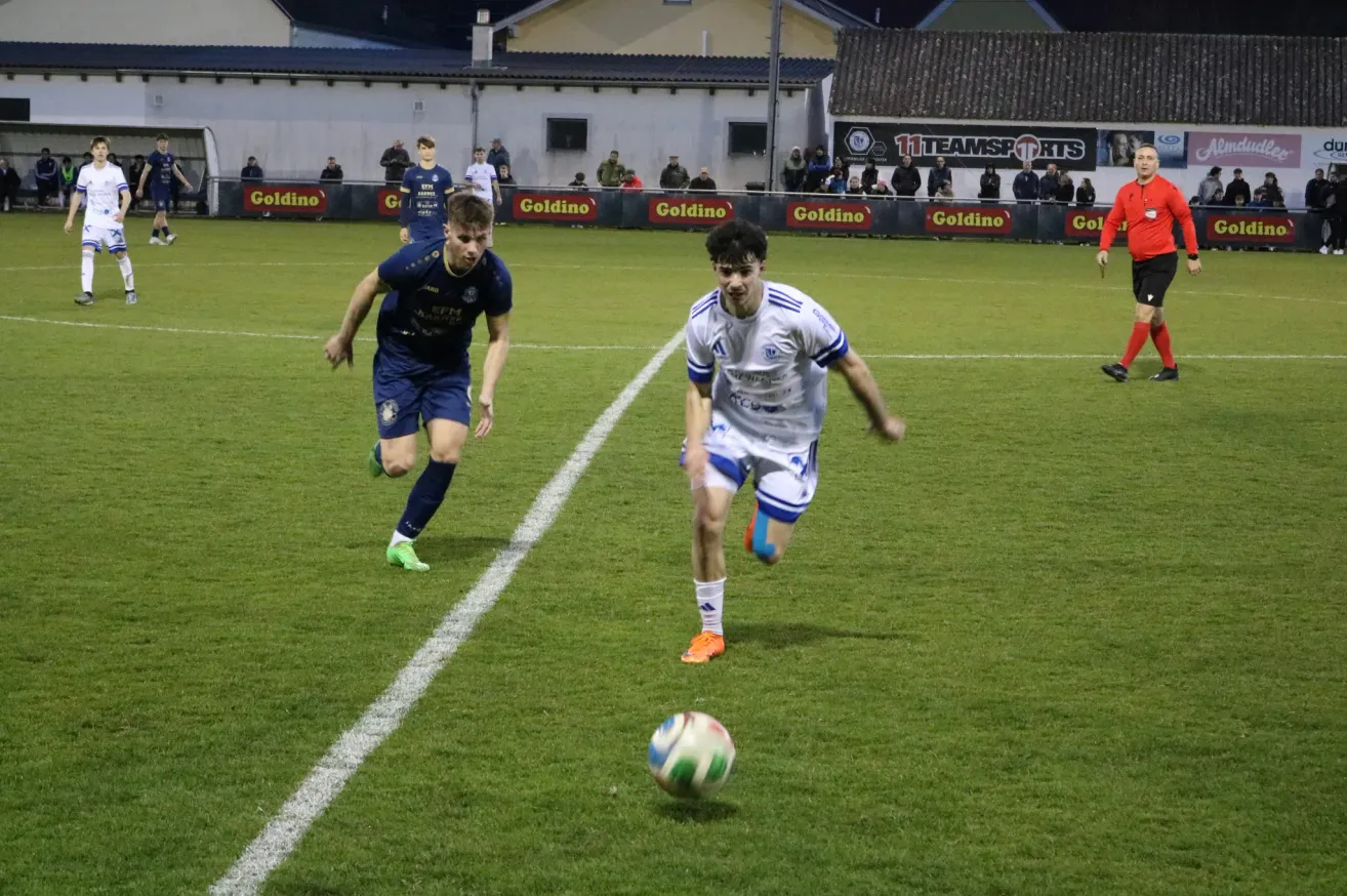Zwei Fußballspieler in blauen und weißen Trikots jagen einem Ball auf einem Rasenfeld nach. Zuschauer sitzen an der Seite und schauen dem Spiel zu. Das Feld ist mit weißen Linien markiert.