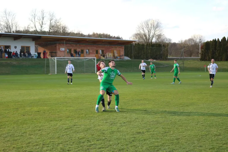 Fußballspieler in grünen Trikots spielen auf einem Feld mit einem Tor. Zuschauer und Spieler sind auf dem Feld und in der Nähe des Gebäudes verteilt.