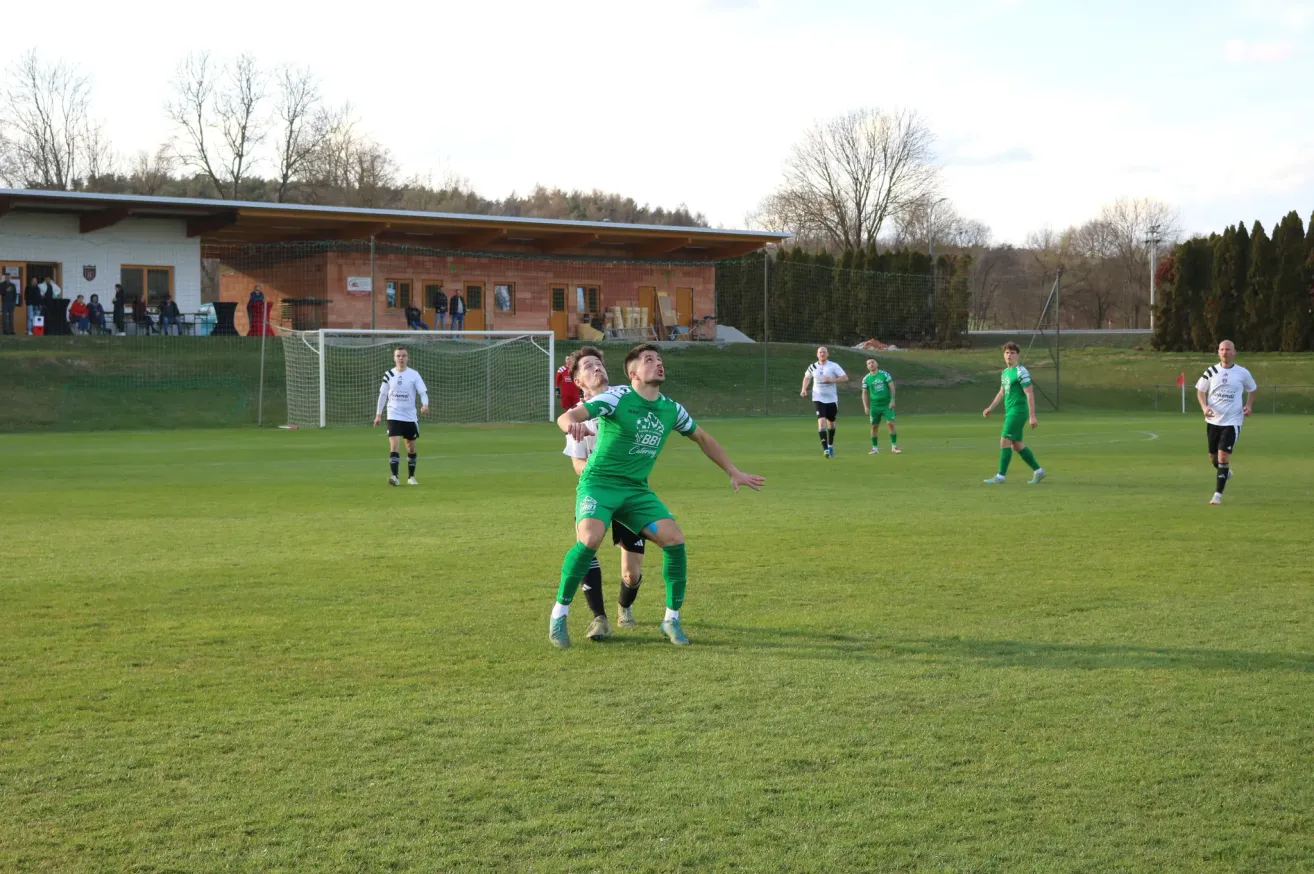 Fußballspieler in grünen Trikots spielen auf einem Feld mit einem Tor. Zuschauer und Spieler sind auf dem Feld und in der Nähe des Gebäudes verteilt.