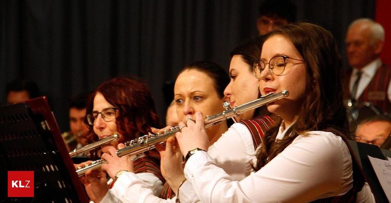 Vier Frauen in einer Band spielen Flöten, wobei eine im Fokus mit einer Brille und einem Armband zu sehen ist. Sie befinden sich in einem Konzertsaal.