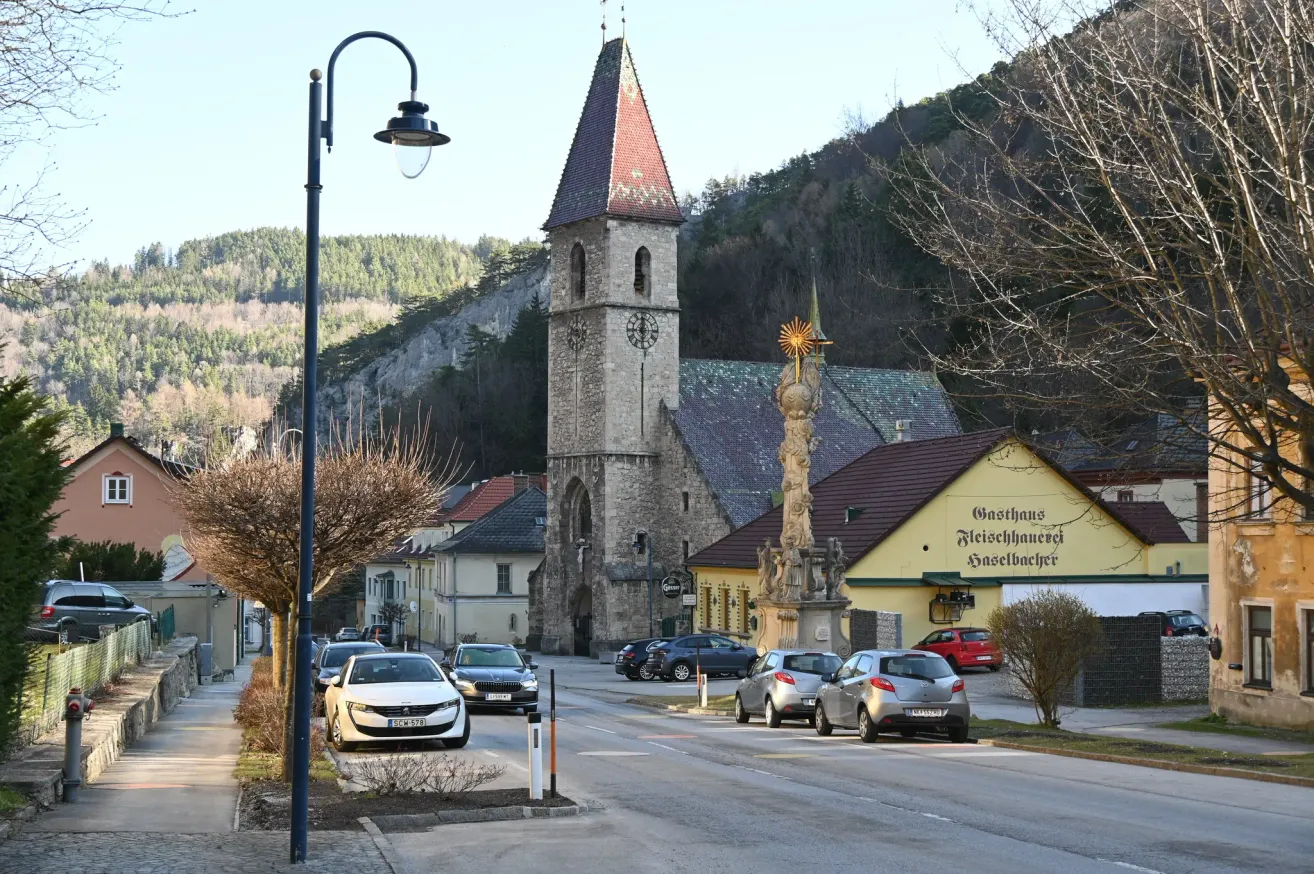Eine Straßenszene mit einem Kirchturm, einer Statue und geparkten Autos auf beiden Seiten. Die Gebäude sind von Bäumen und Bergen umgeben.
