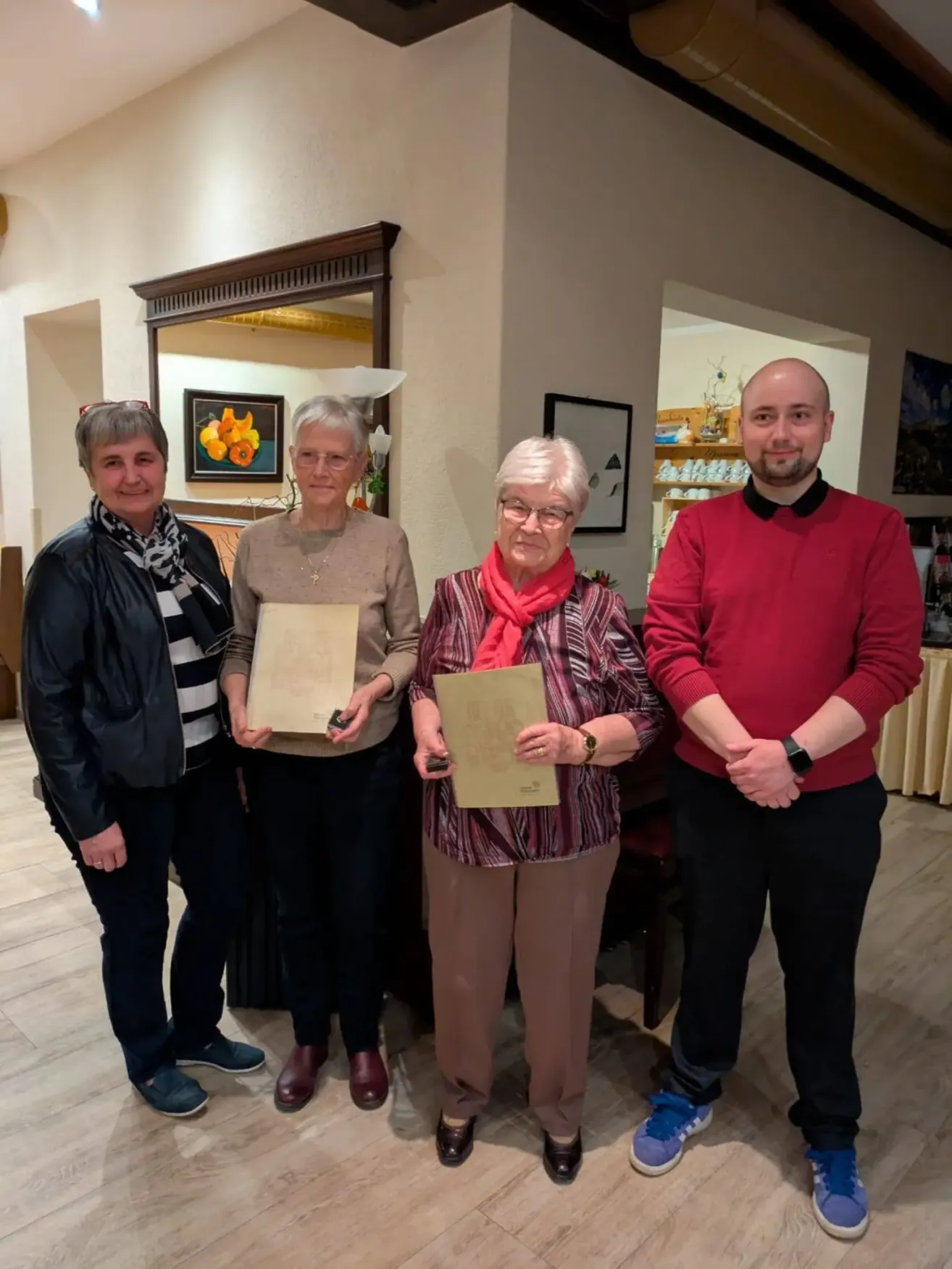 Vier Menschen stehen drinnen und halten Urkunden. Eine Frau links trägt ein Tuch, eine andere Frau in der Mitte trägt eine Brille. Der Mann rechts hat einen kahlen Kopf und trägt ein rotes Hemd. Hinter ihnen befindet sich ein Spiegel mit Rahmen und ein Regal mit Gegenständen.