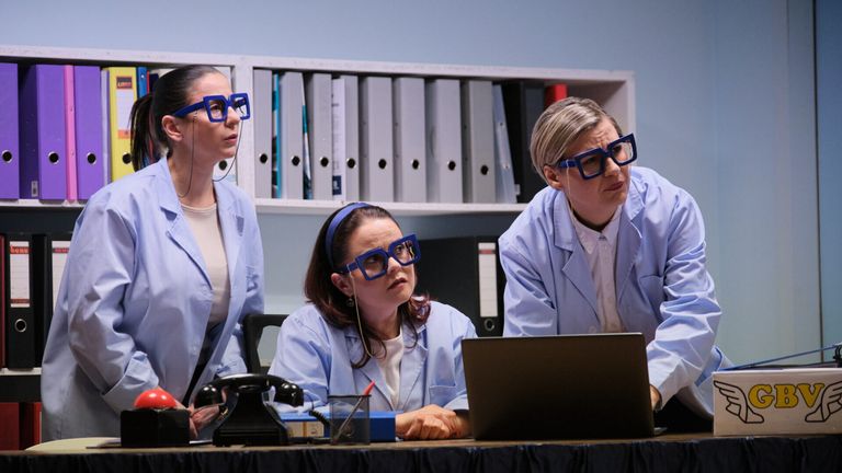 Drei Frauen in Laborkittel mit großen blauen Gläsern sitzen um einen Schreibtisch herum. Die Frau in der Mitte schaut auf einen Laptop, während die anderen beiden auf etwas außerhalb des Bildes konzentriert zu sein scheinen. Ein Telefon und eine Box stehen auf dem Schreibtisch.