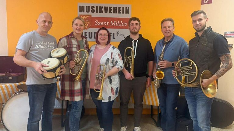 Fünf Personen posieren für ein Foto, jeder hält Musikinstrumente, vor einer gelben Wand mit einem Schild, das 'Musikverein Szent Miklos' steht.