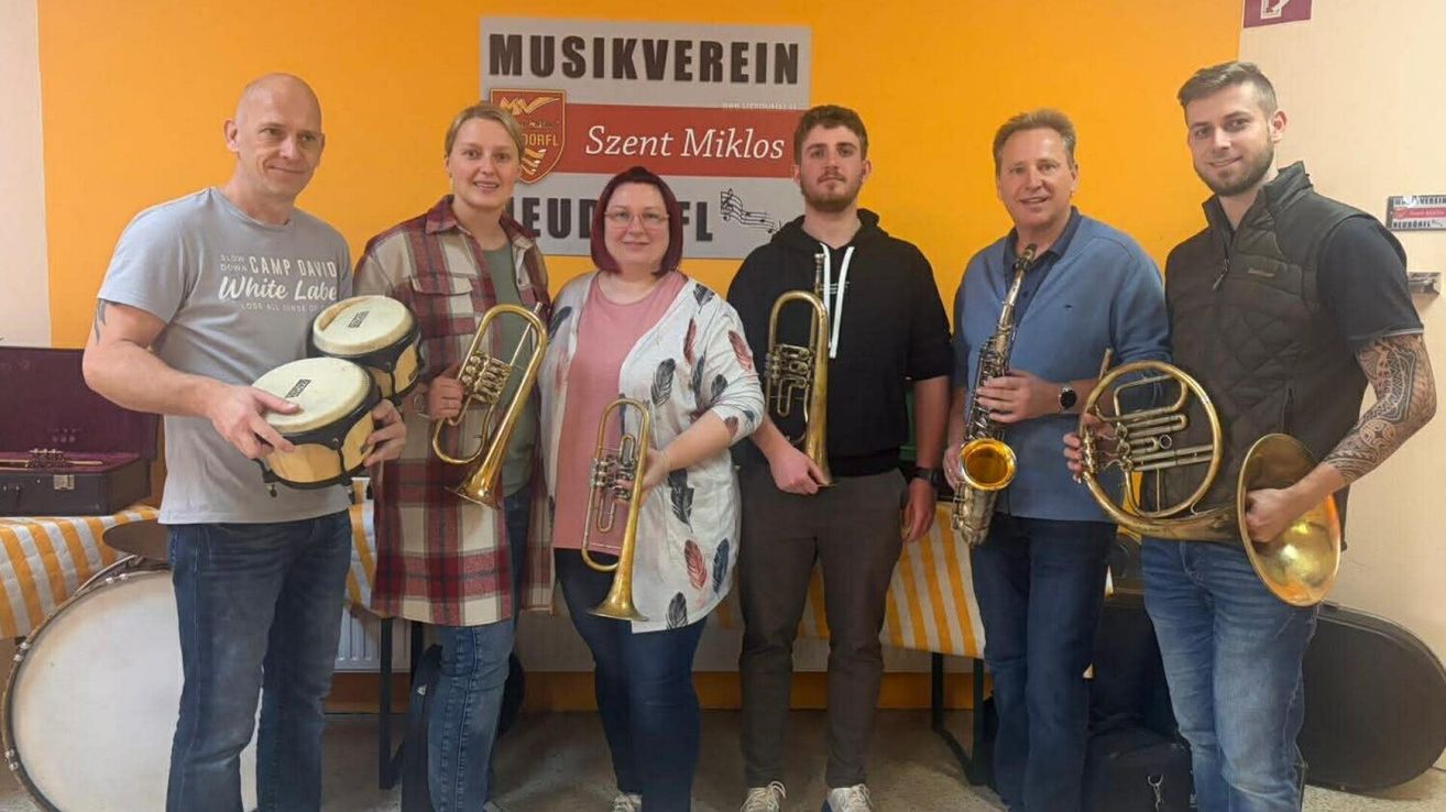 Fünf Personen posieren für ein Foto, jeder hält Musikinstrumente, vor einer gelben Wand mit einem Schild, das 'Musikverein Szent Miklos' steht.