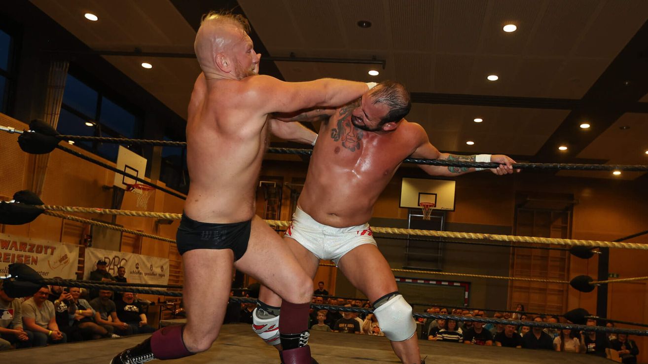 Zwei Wrestler befinden sich im Ring. Einer hält den Arm und das Bein des anderen fest, während der andere seinen Kopf festhält. Das Publikum beobachtet sie von den Tribünen.