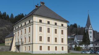 Ein großes Gebäude mit einem Schieferdach und vielen Fenstern. Das Gebäude ist von einem grünen Rasen umgeben. In der Ferne befinden sich weitere Gebäude und Bäume.