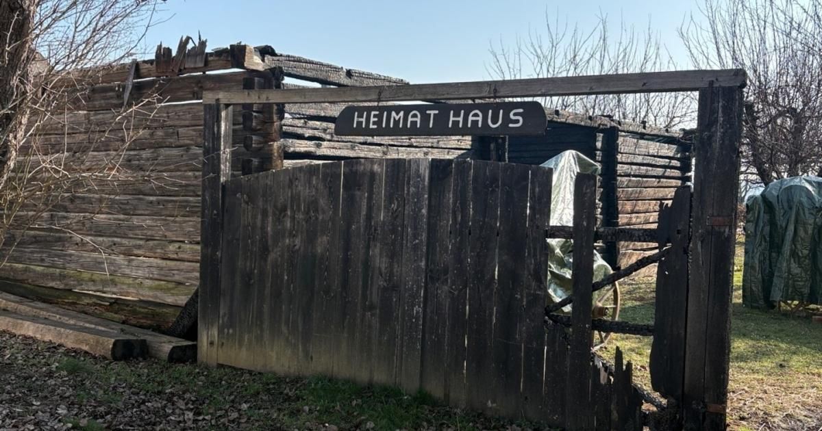 Ein hölzerne Tür mit einem Schild, das 'Heimat Haus' besagt. Das Tor ist alt und verwittert, neben einem Holzschuppen gelegen.