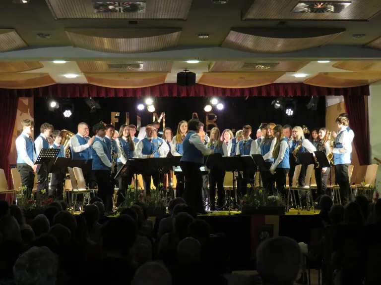 Eine Band spielt auf der Bühne vor einem Publikum. Die Bühne hat einen roten Vorhang als Hintergrund mit den Buchstaben FIM. Die Musiker tragen blaue Uniformen.