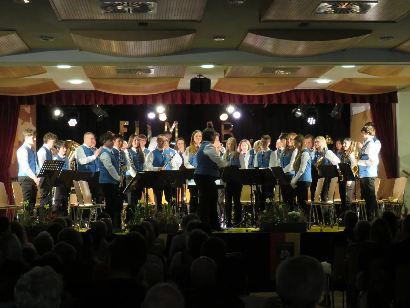 Eine Band spielt auf der Bühne vor einem Publikum. Die Bühne hat einen roten Vorhang als Hintergrund mit den Buchstaben FIM. Die Musiker tragen blaue Uniformen.