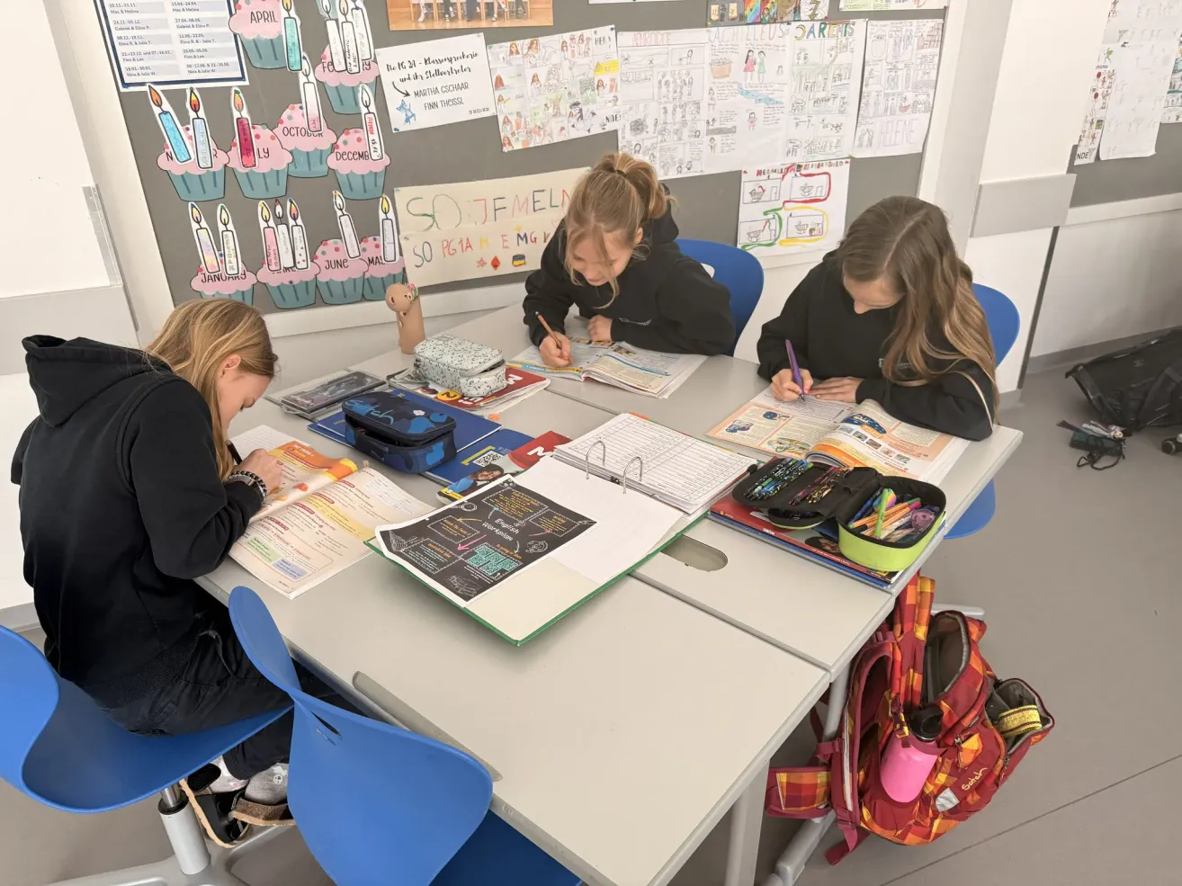 Drei junge Mädchen sitzen in einem Klassenzimmer an einem Schreibtisch und schreiben mit Stiften auf Papier. Der Schreibtisch ist mit Notizbüchern, Stiften und anderen Schulsachen gefüllt. Hinter ihnen ist eine Wand mit Zeichnungen und einem Kalender.