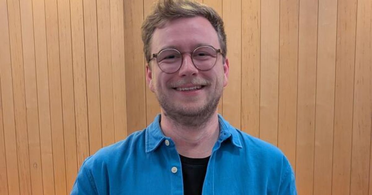 Ein Mann mit Brille und einem blauen Hemd mit schwarzem Innenhemd lächelt vor einer Holzwand.